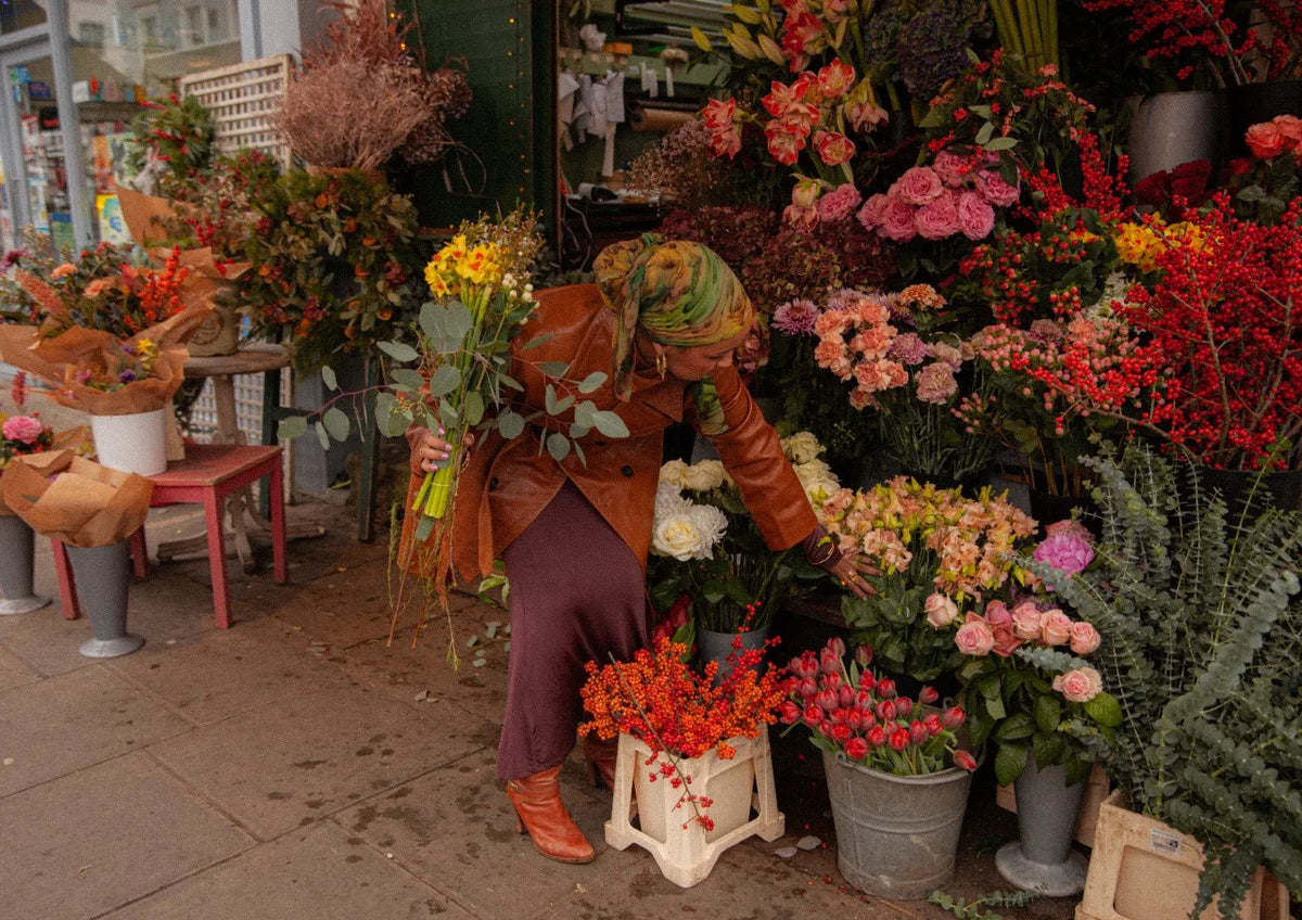 Frau im braunen Ledermantel stellt an einem Blumenstand auf dem Gehweg einen Strauß zusammen